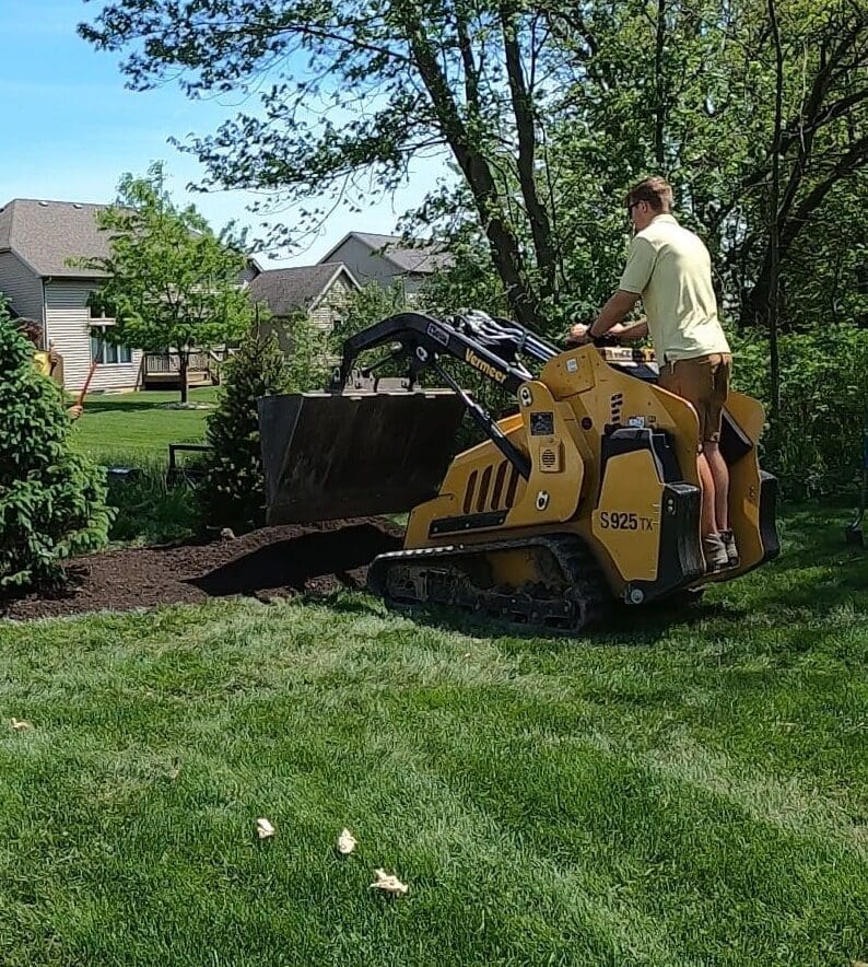 Fort Wayne landscaper using compact track loader to spread mulch in suburban yard.
