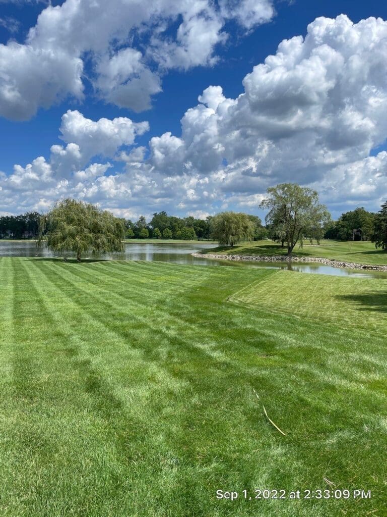 Freshly mowed Fort Wayne park lawn beside calm lake under blue sky and clouds.