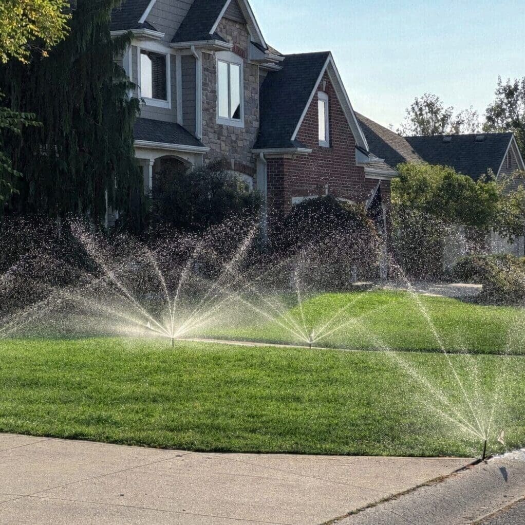 Fort Wayne irrigation system sprinklers watering a green suburban front lawn in sunlight.